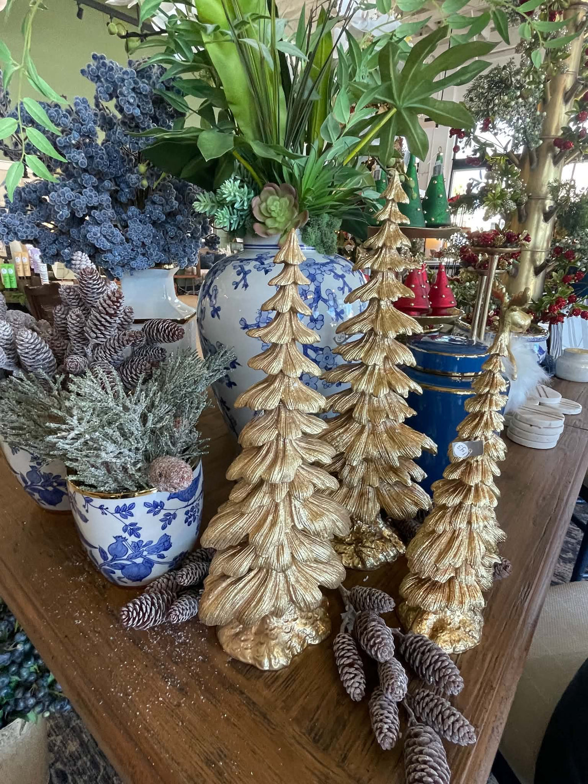 Three metallic gold Christmas tree ornaments displayed on a wooden table alongside blue and white ceramic pots, faux pine trees, and decorative pinecones.
