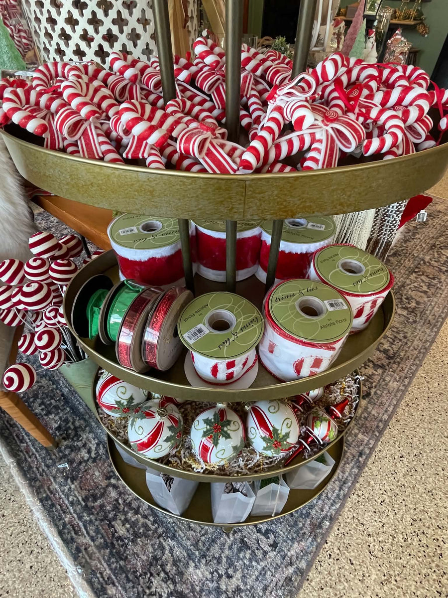 A gold-colored three-tier display stand filled with holiday decorations, including red and white striped candy canes on the top shelf, spools of red and white ribbon on the middle shelf, and white ornaments with red and green holiday designs on the bottom shelf.