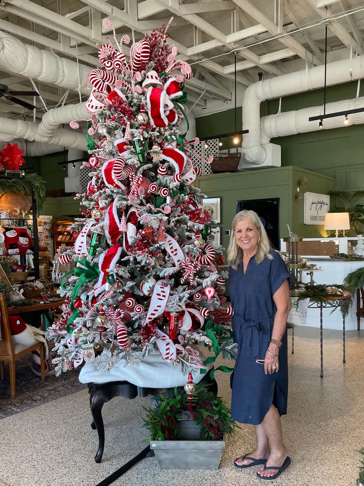 A woman in a navy blue dress stands next to a large, decorated Christmas tree in a retail store setting. The tree is adorned with red and white candy cane ribbons, white and red striped ornaments, and green pine branches.