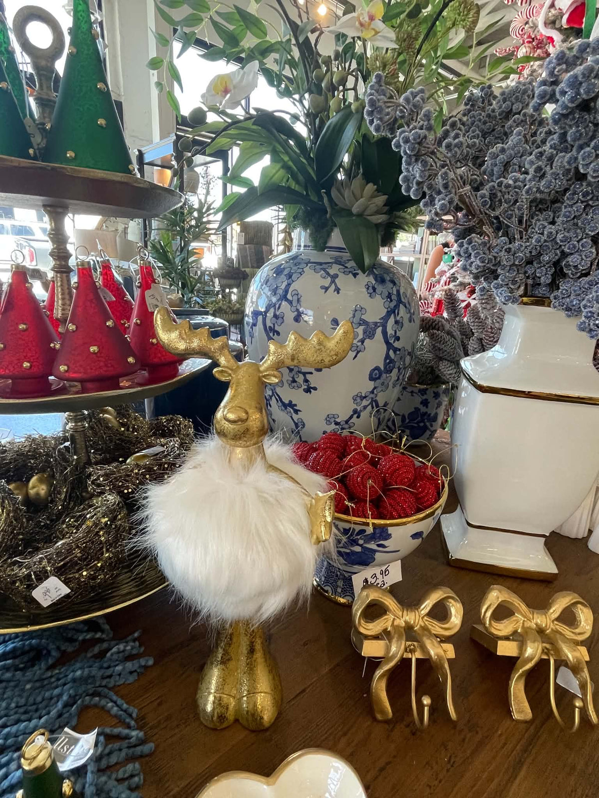 A festive holiday display on a wooden table featuring a gold reindeer figurine with a white faux-fur neck, red Christmas tree ornaments on a tiered stand, a blue and white porcelain vase with flowers, and gold bow decorations.