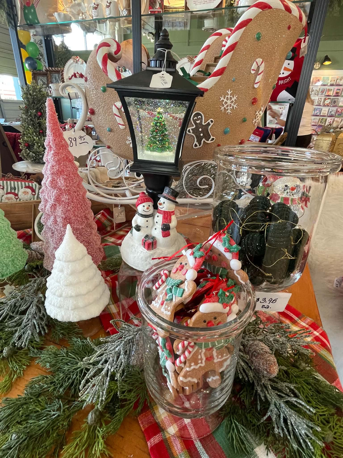 A festive holiday display featuring a clear glass jar filled with gingerbread cookies, surrounded by miniature Christmas trees, a snowman figurine, a lantern, and other seasonal decorations on a table.