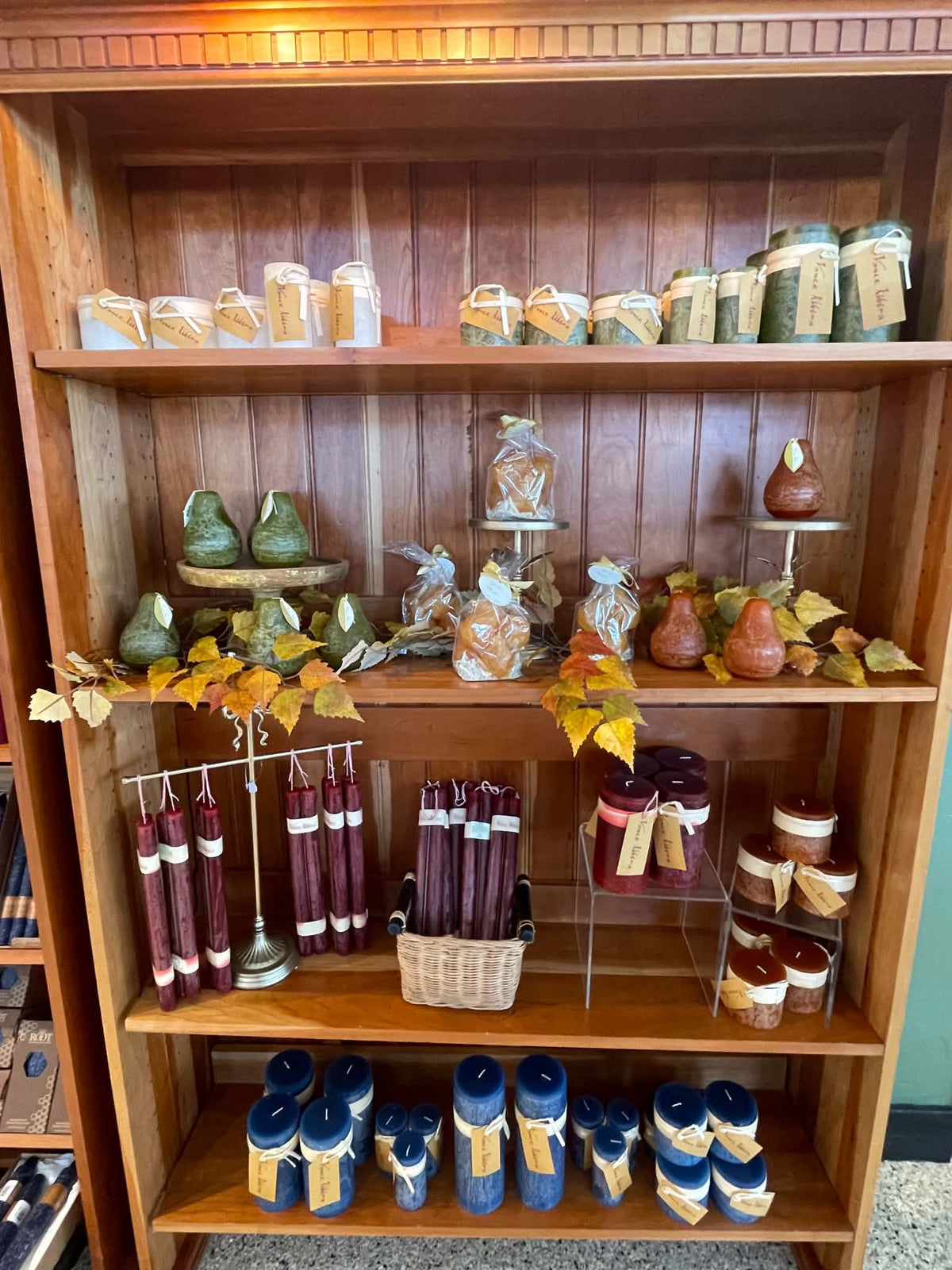 A wooden display shelf filled with assorted home goods and candles, including blue and burgundy wax candles, small jars, decorative gourds, and fall-themed decor.