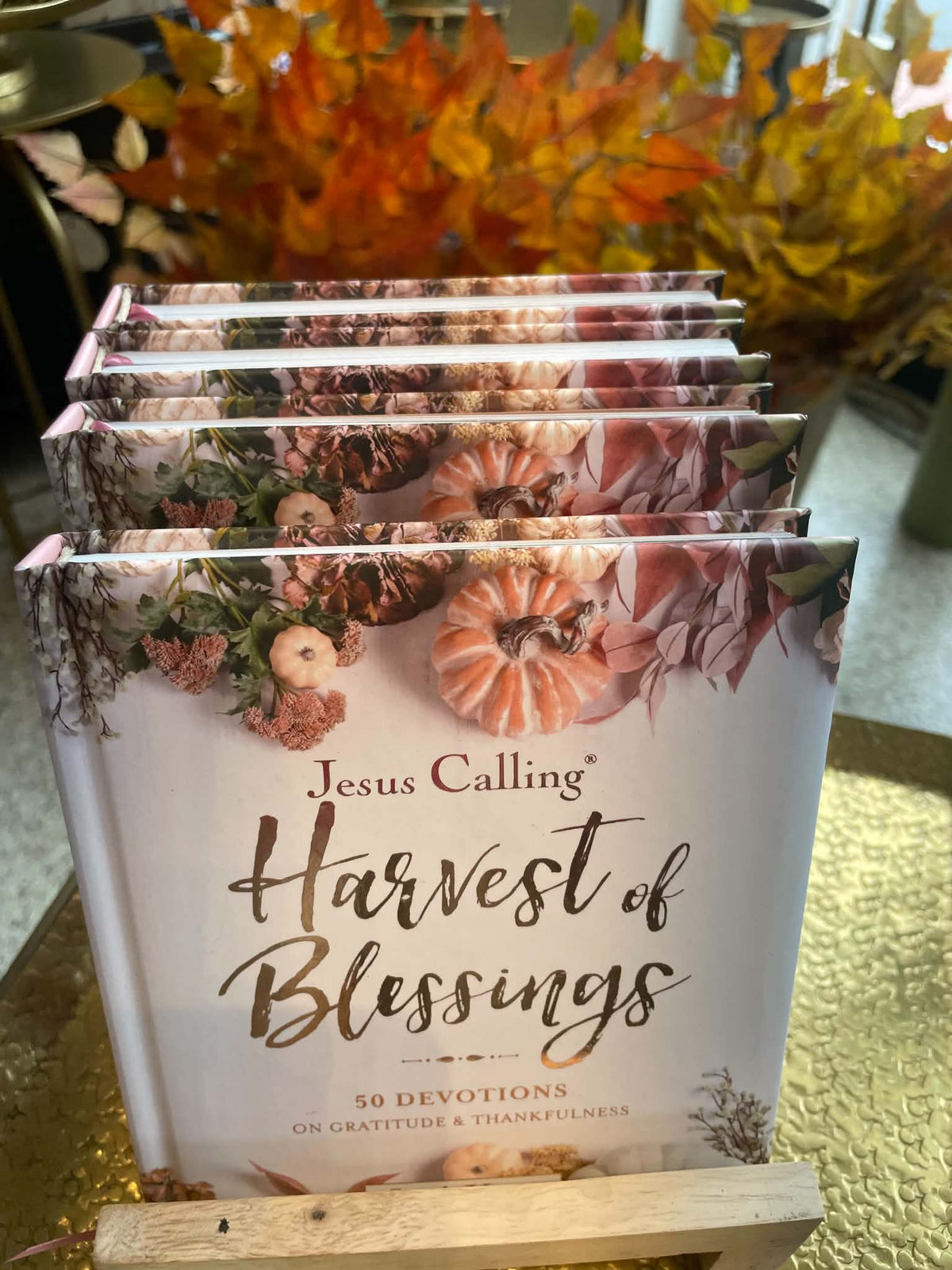 A stack of 'Jesus Calling Harvest of Blessings' devotional books displayed on a small wooden stand, with autumn foliage in the background.