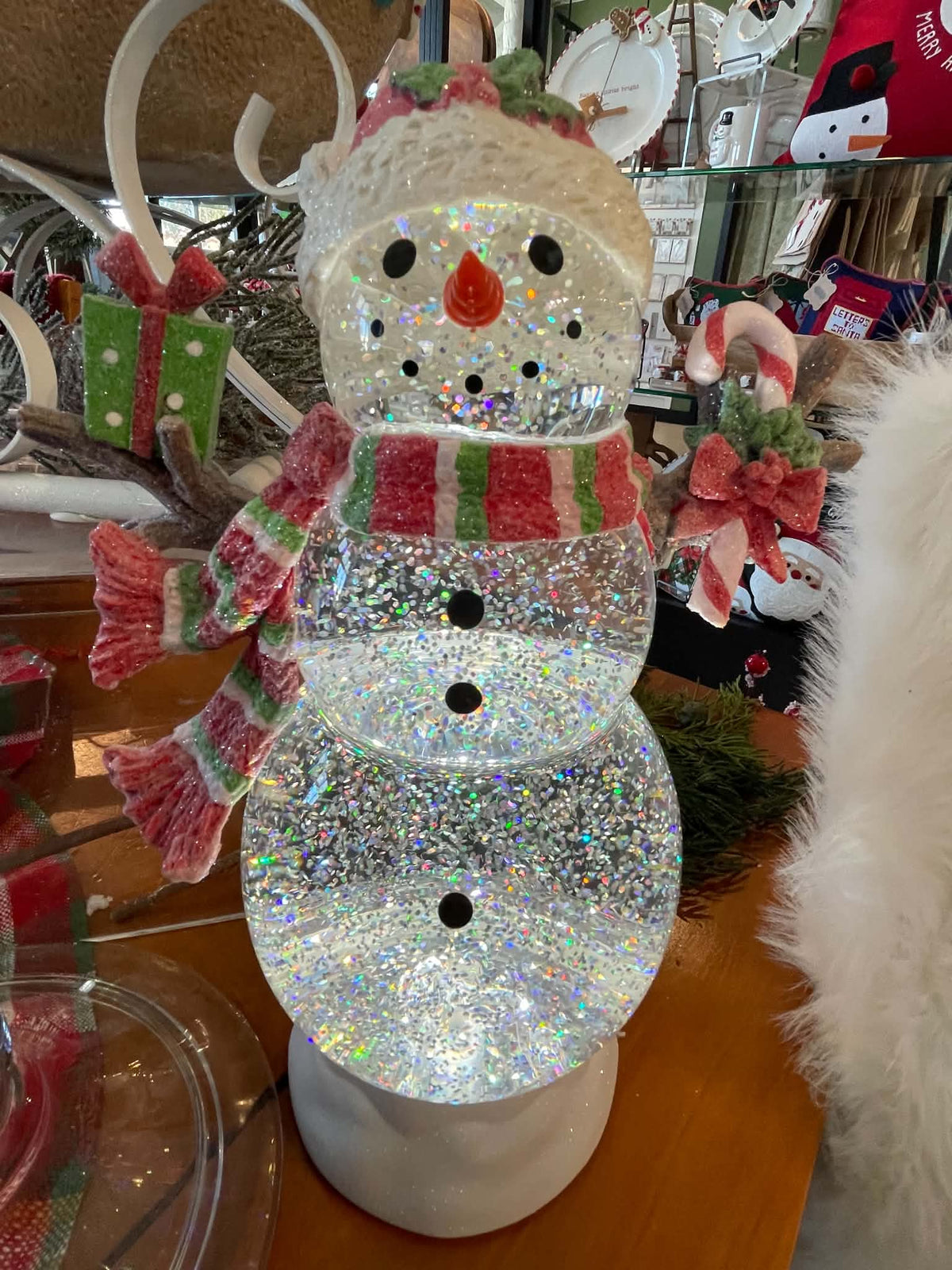 A glittery snowman Christmas decoration with a white knit hat, red and green striped scarf, and black button details, displayed on a wooden surface in a store setting.