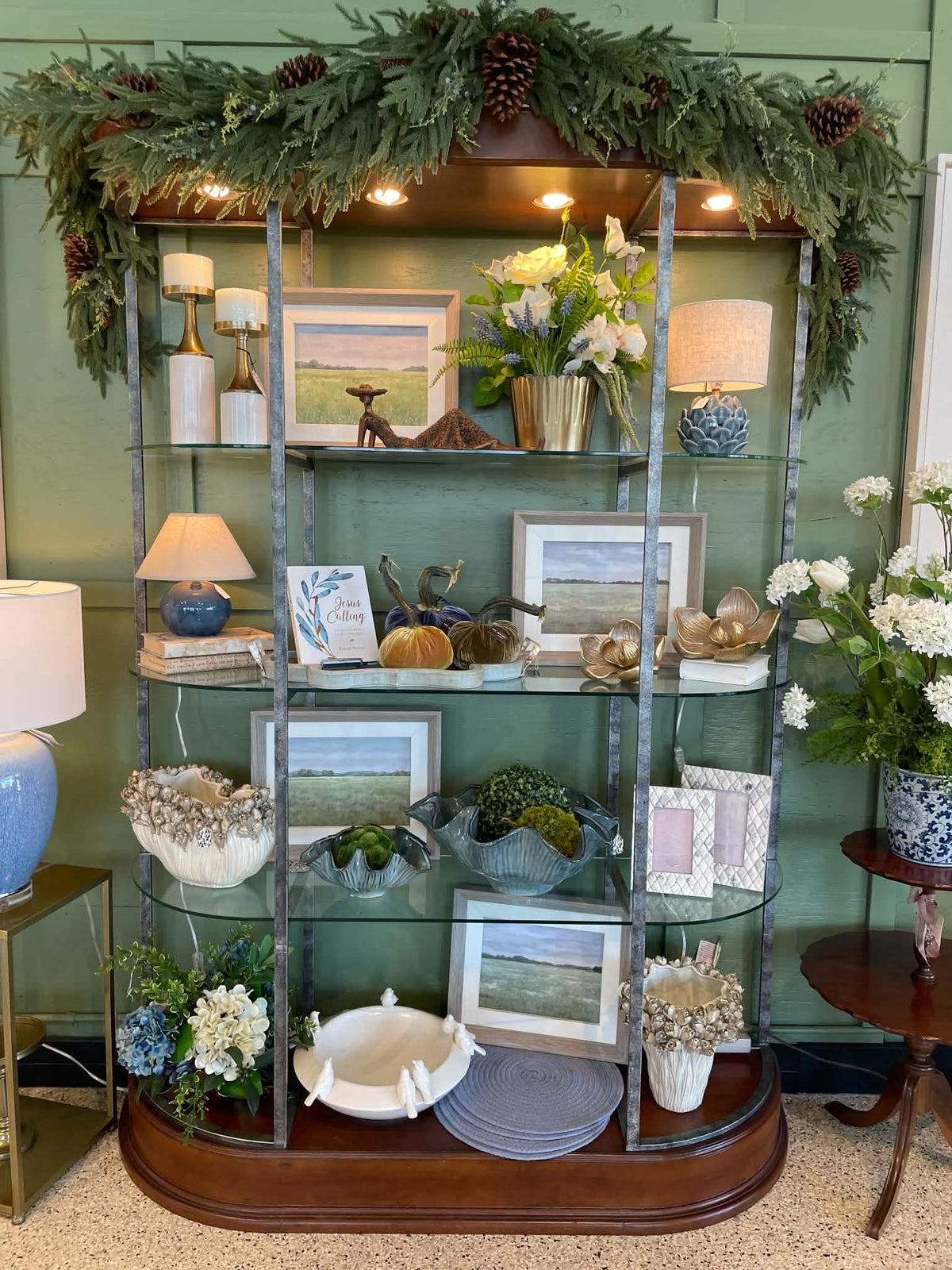 A large, ornate wooden display bookcase with glass shelves, styled with home decor items including framed prints, lamps, flowers, and a green pine garland with pinecones across the top, set against a green paneled wall.