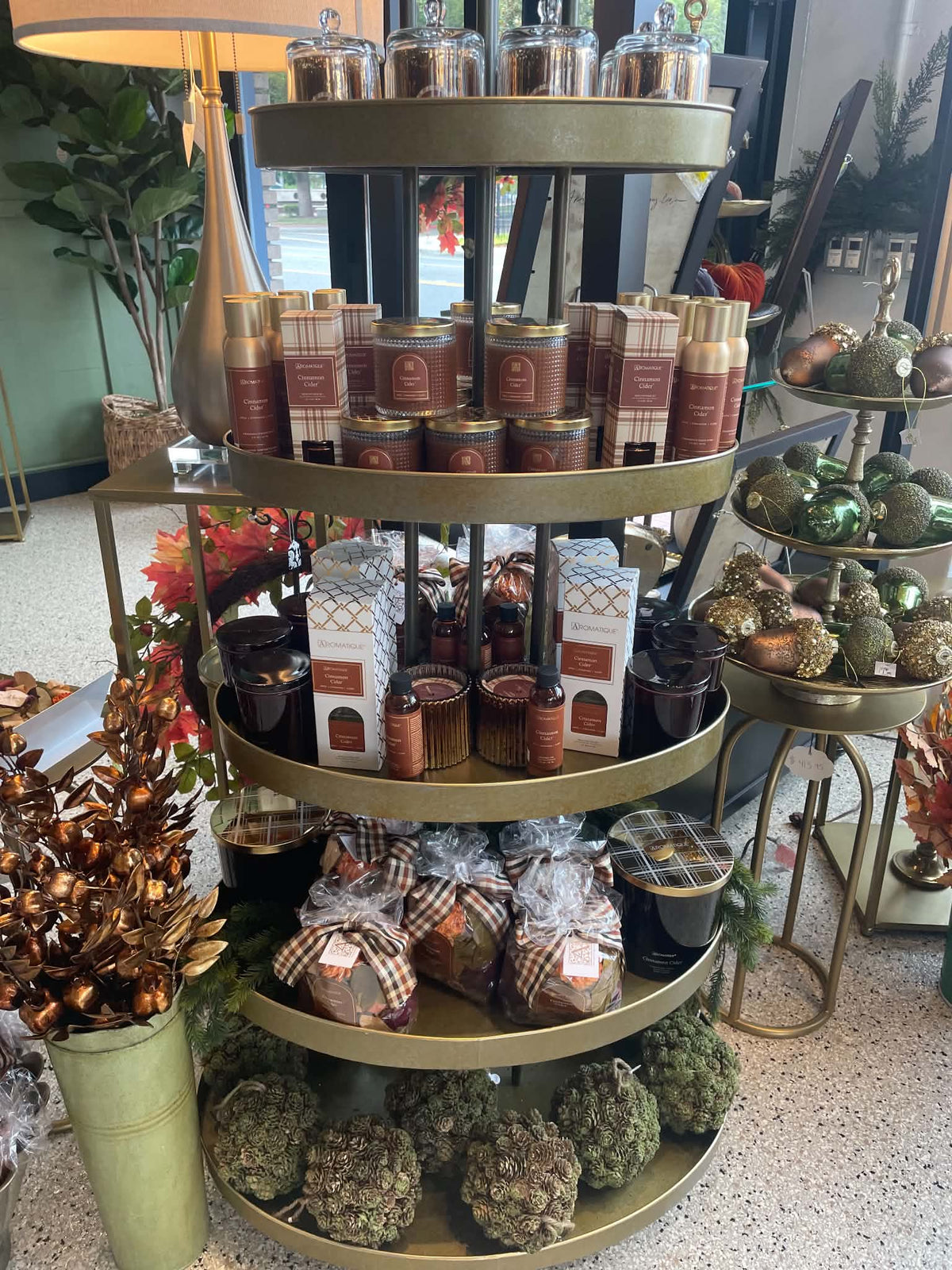 A three-tier gold rolling display cart filled with assorted home fragrance and gift items, including candles, lotions, and packaged scented products, set in a retail store environment.