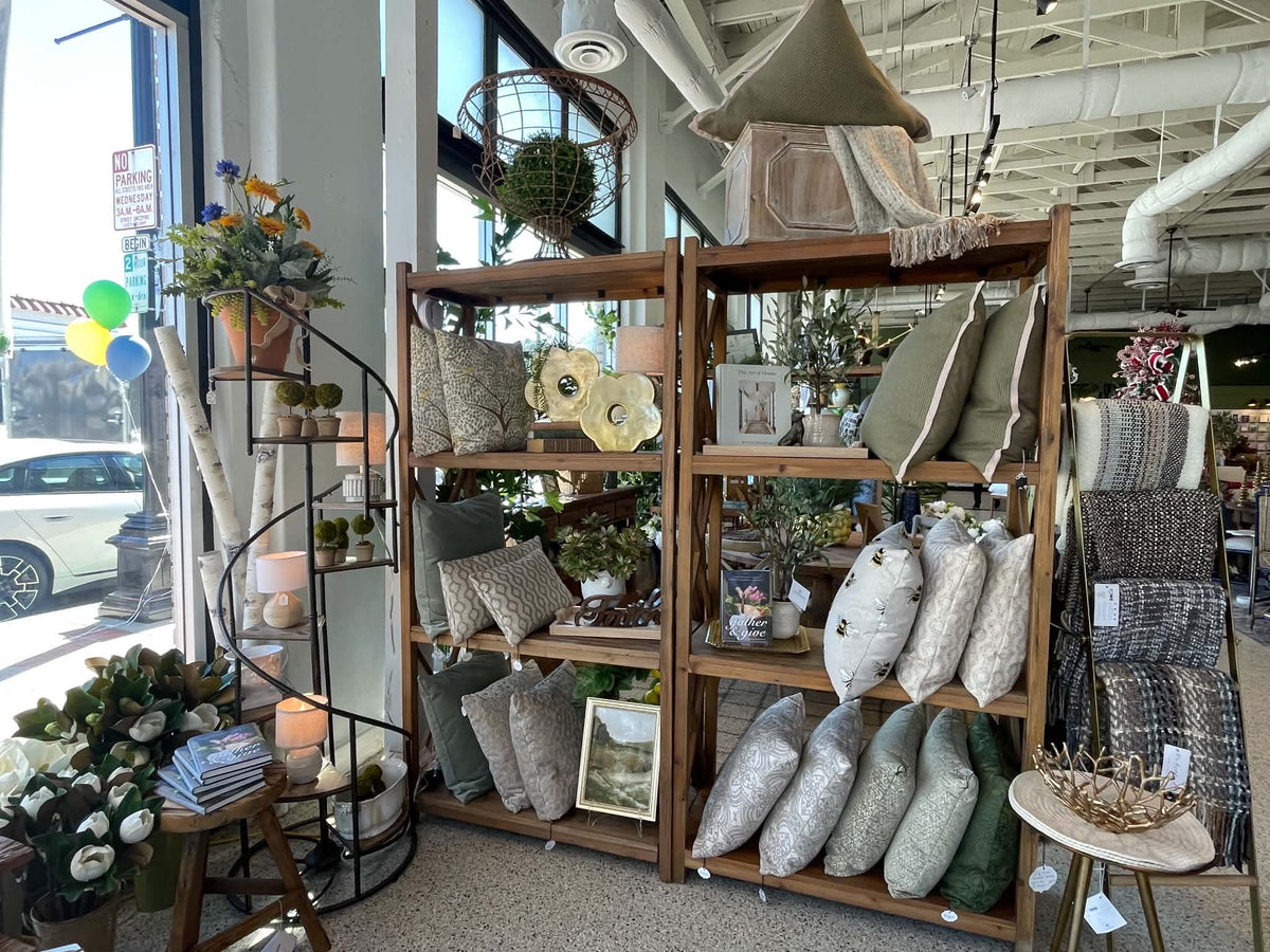 A rustic wooden display shelf filled with decorative throw pillows, plants, and home decor items inside a retail store. The shelf is surrounded by other home decor products, a spiral staircase, and large windows showing a street scene outside.