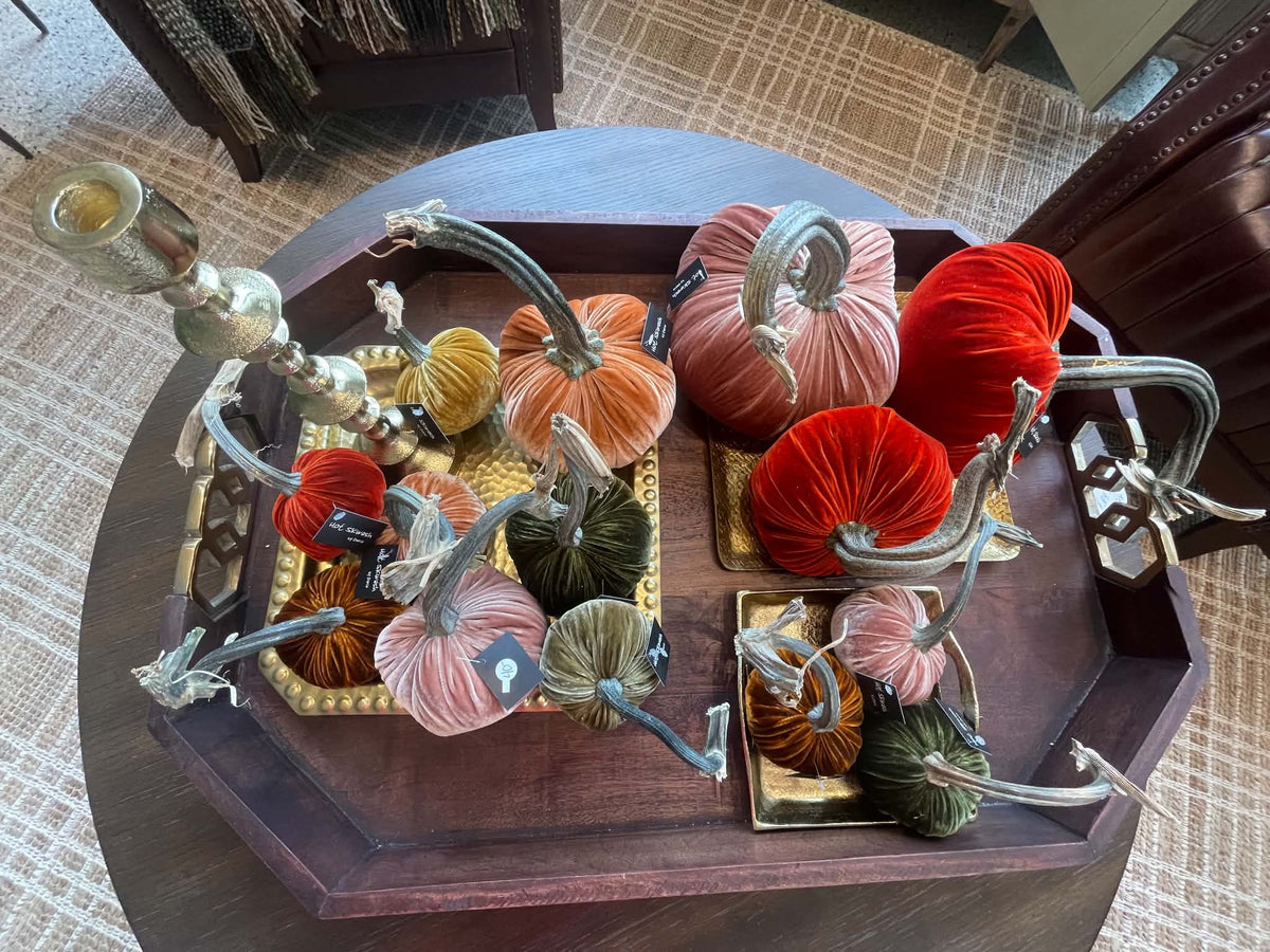 A decorative tabletop arrangement of velvet pumpkins in red, pink, orange, green, and mustard tones, styled on a dark wood octagonal tray with silver-toned metal stems and candleholders.
