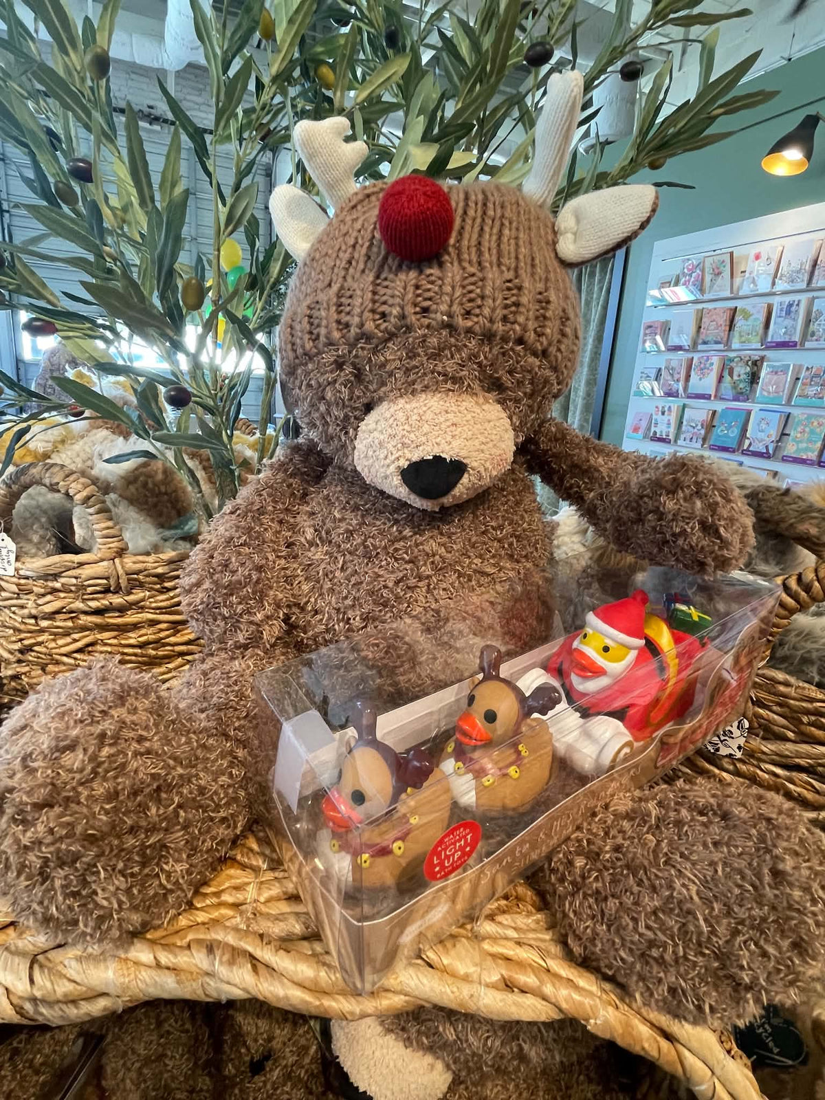 A brown plush teddy bear wearing a knitted reindeer hat with white antlers and a red nose, sitting on a wicker basket display. In front of the bear is a clear plastic box containing small holiday-themed plush figures.
