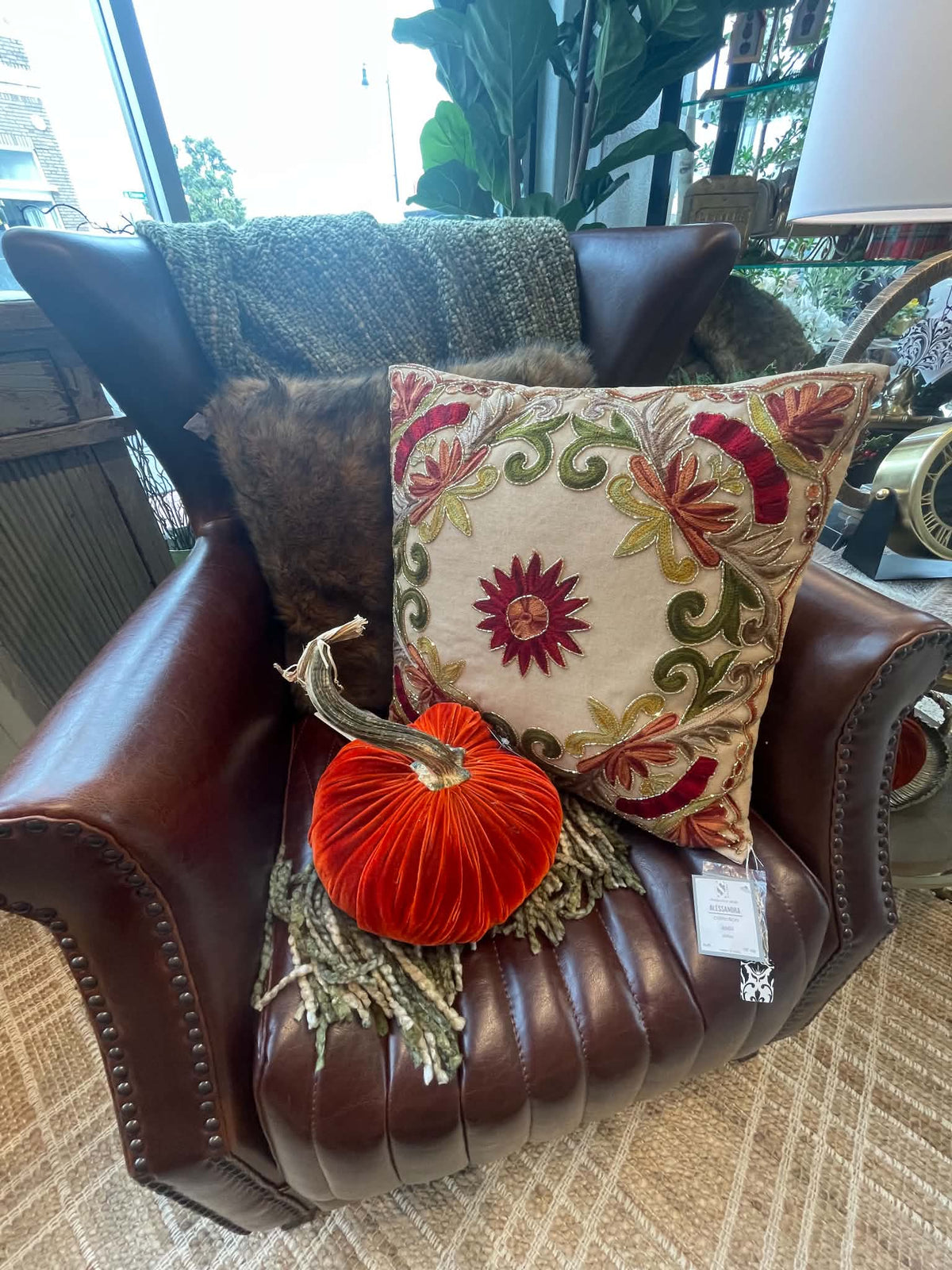 A brown leather armchair in a home setting, styled with a cream floral throw pillow, a red velvet pumpkin decoration, and a dark knit blanket.