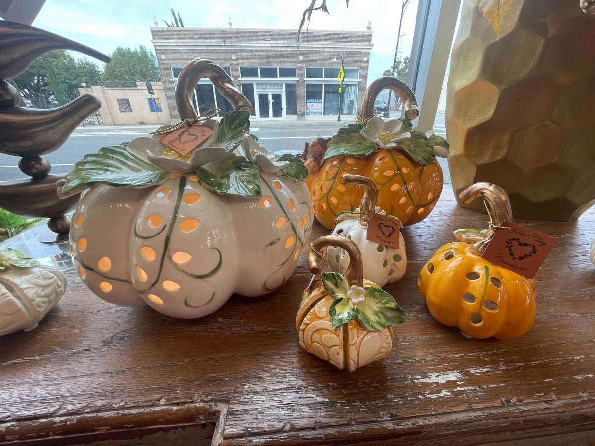 A group of decorative ceramic pumpkins displayed on a wooden shelf. The set includes several pumpkins in cream, orange, and white glazes, with green leaf details, carved cutout windows showing warm light from within, and small brown tags attached.