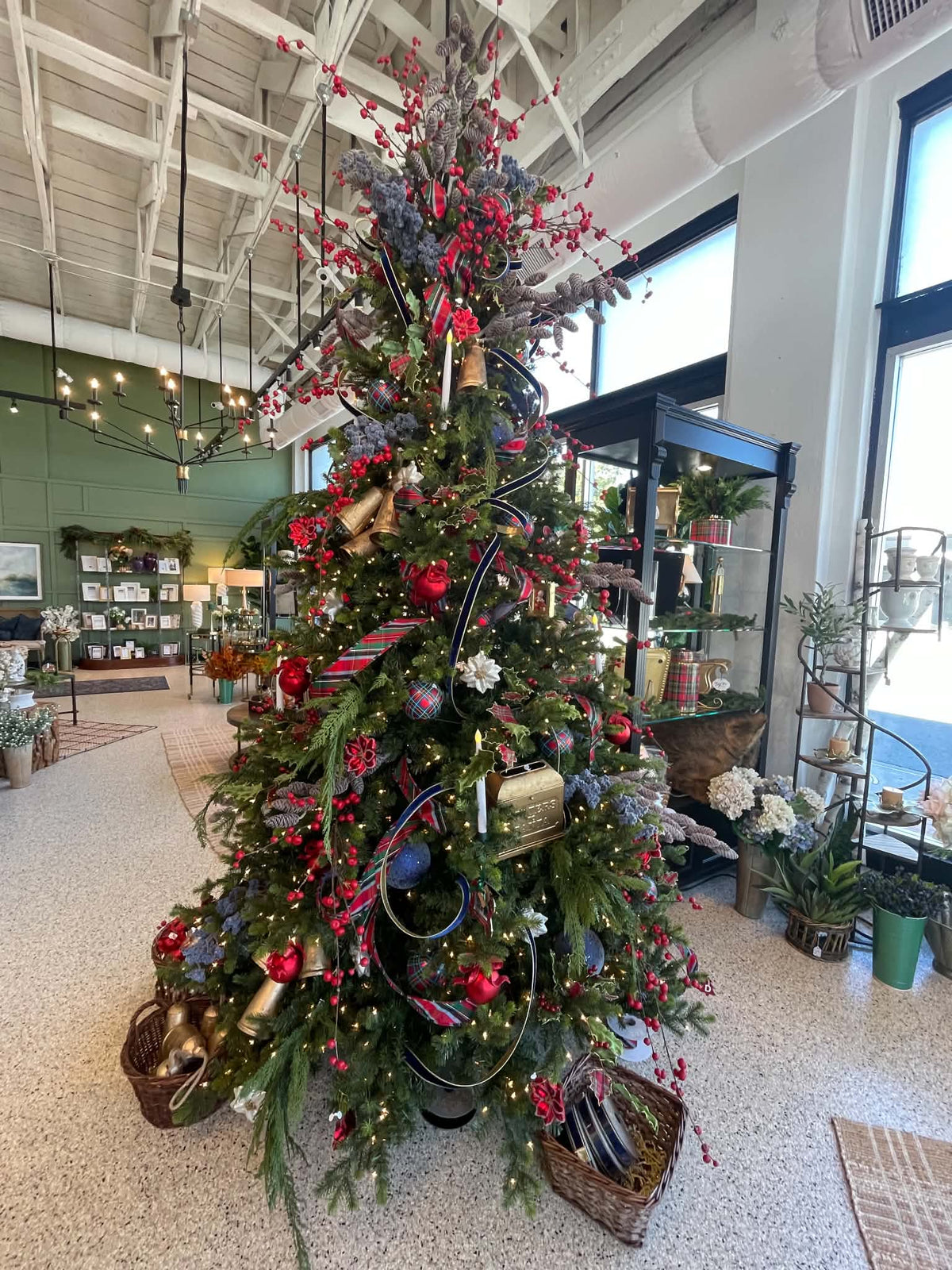 A decorated Christmas tree in a retail shop interior, adorned with red berries, gold and silver ornaments, ribbons, and wicker baskets on a speckled floor.