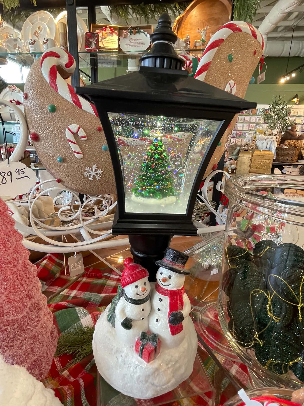 A black lantern-shaped snow globe decoration with a miniature Christmas tree inside, displayed on a table with holiday decorations including gingerbread cookies, candy canes, and snowman figurines.