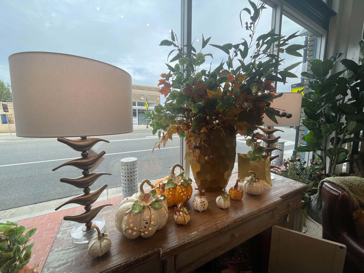 A styled autumn table arrangement featuring a large beige table lamp, a gold ceramic vase filled with green foliage and fall leaves, and several decorative pumpkins in white, orange, and beige on a wooden console table near a window.