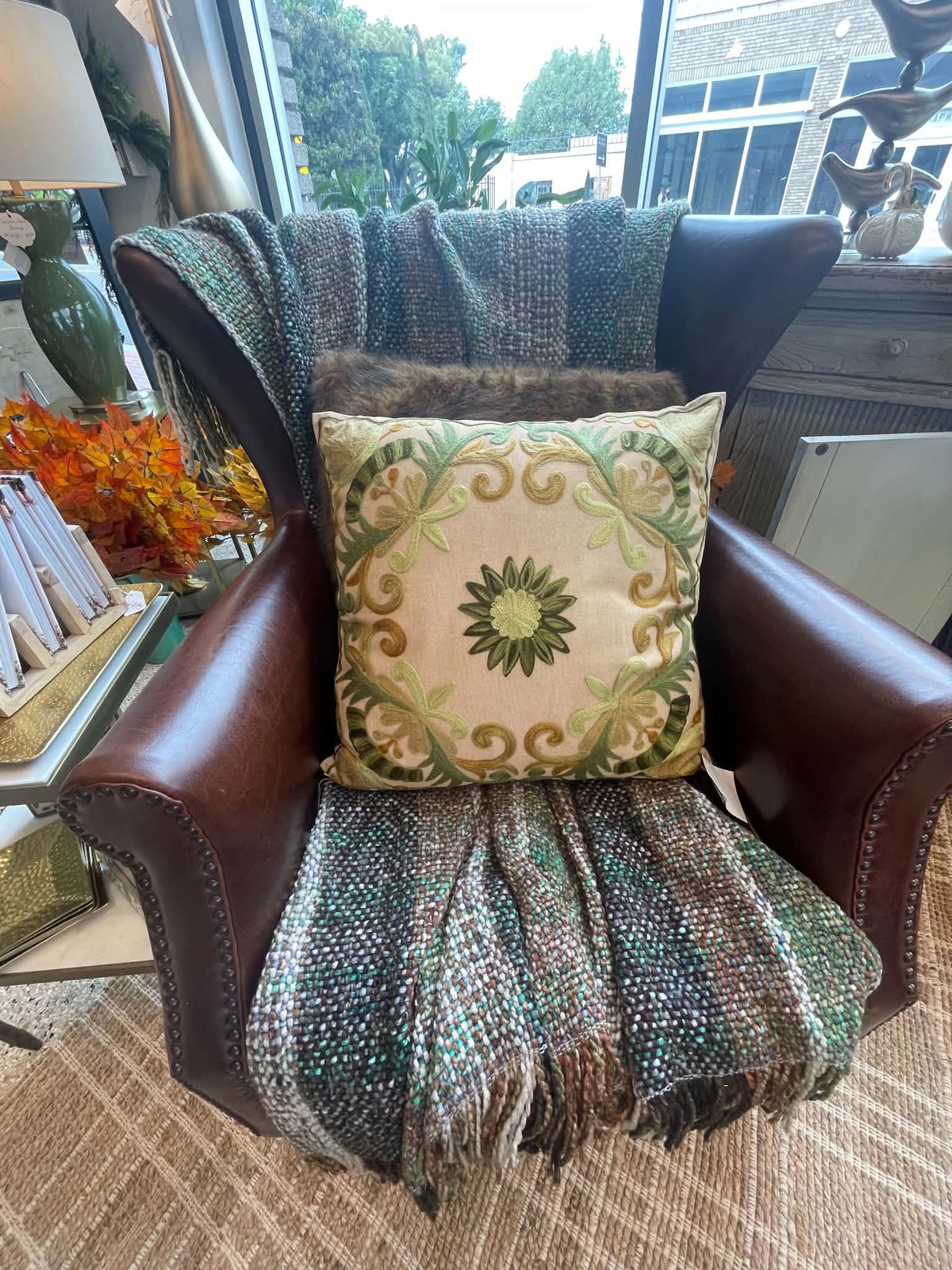 A square decorative throw pillow with a green and beige floral scroll pattern, styled on a brown leather wingback armchair draped with a green and brown woven blanket.