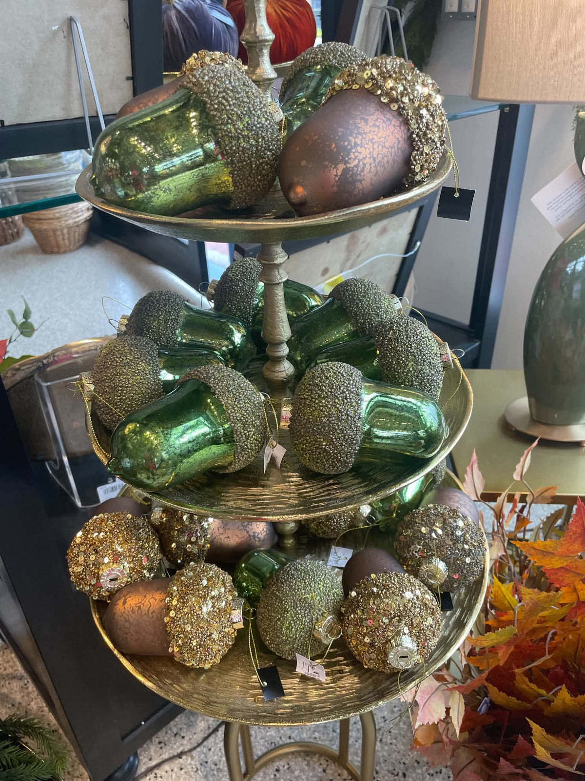 Three-tier gold metal display stand filled with decorative acorns and acorn-like ornaments in metallic green, bronze, and glittery gold, photographed in a store interior.